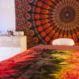 Czas Relaksu Tomasz Marciniak - Przytulne wnętrze gabinetu z łóżkiem do masażu przykrytym barwnym materiałem tie-dye, w tle mandala na ścianie i półka z buteleczkami olejków eterycznych.