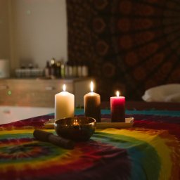 Czas Relaksu Tomasz Marciniak - Przytulne wnętrze gabinetu z łóżkiem do zabiegów, na którym leży kolorowy pled w stylu tie-dye, a na nim ustawione trzy zapalone świece o różnych kolorach i miska tybetańska z pałeczką; w tle...