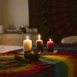 Przytulne wnętrze gabinetu z łóżkiem do zabiegów, na którym leży kolorowy pled w stylu tie-dye, a na nim ustawione trzy zapalone świece o różnych kolorach i miska tybetańska z pałeczką; w tle...