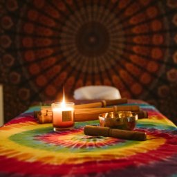 Czas Relaksu Tomasz Marciniak - Stół do masażu przykryty kolorową tkaniną w stylu tie-dye, na nim świeca, miseczka dźwiękowa i bambusowe pałeczki, w tle dekoracyjna mandala na ścianie.