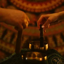 Dwie dłonie wprawiają w ruch misy tybetańskie na tle wzorzystej tkaniny. Ujęcie z bliska, ciepłe światło, relaksacyjny klimat.