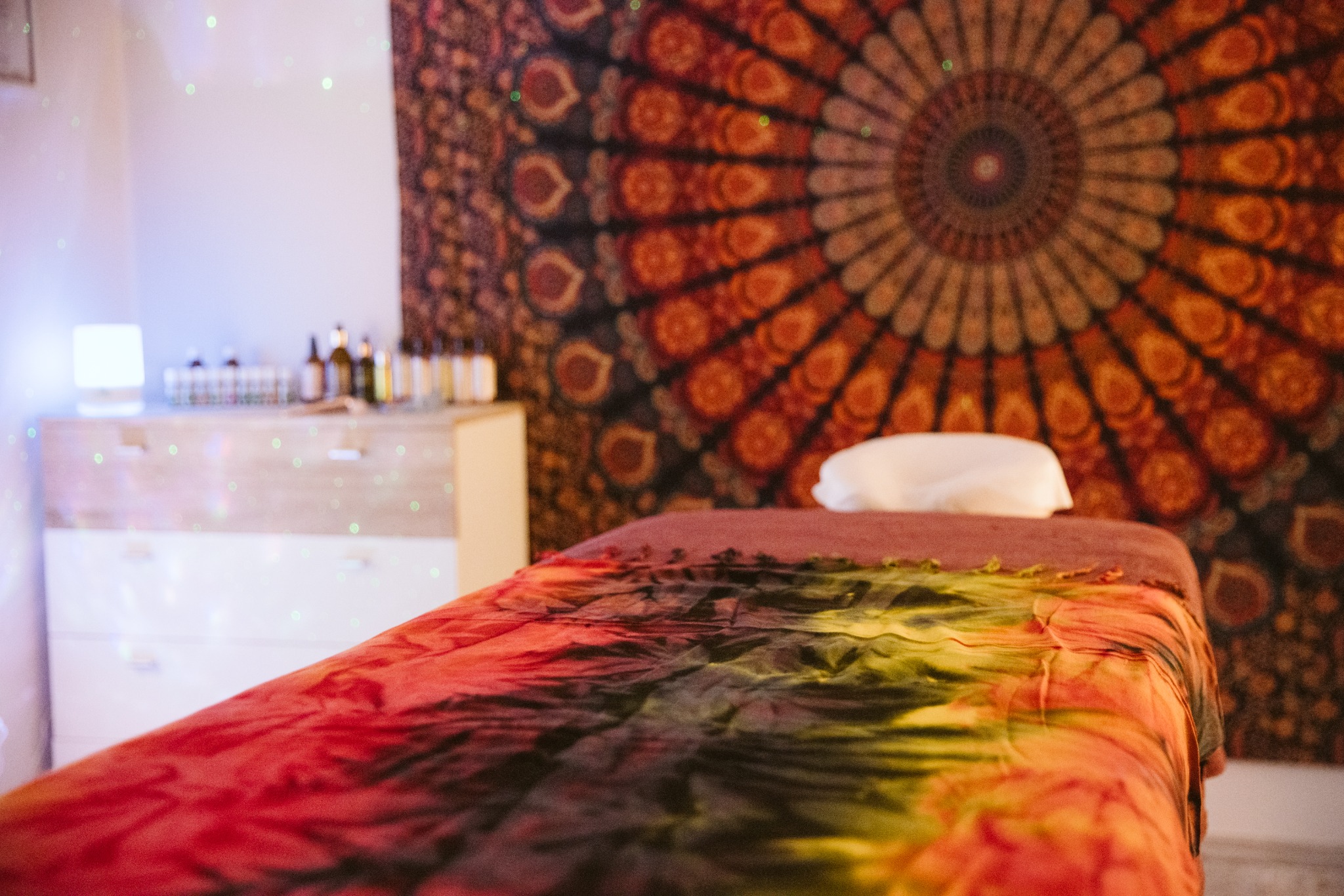Przytulne wnętrze gabinetu z łóżkiem do masażu przykrytym barwnym materiałem tie-dye, w tle mandala na ścianie i półka z buteleczkami olejków eterycznych.