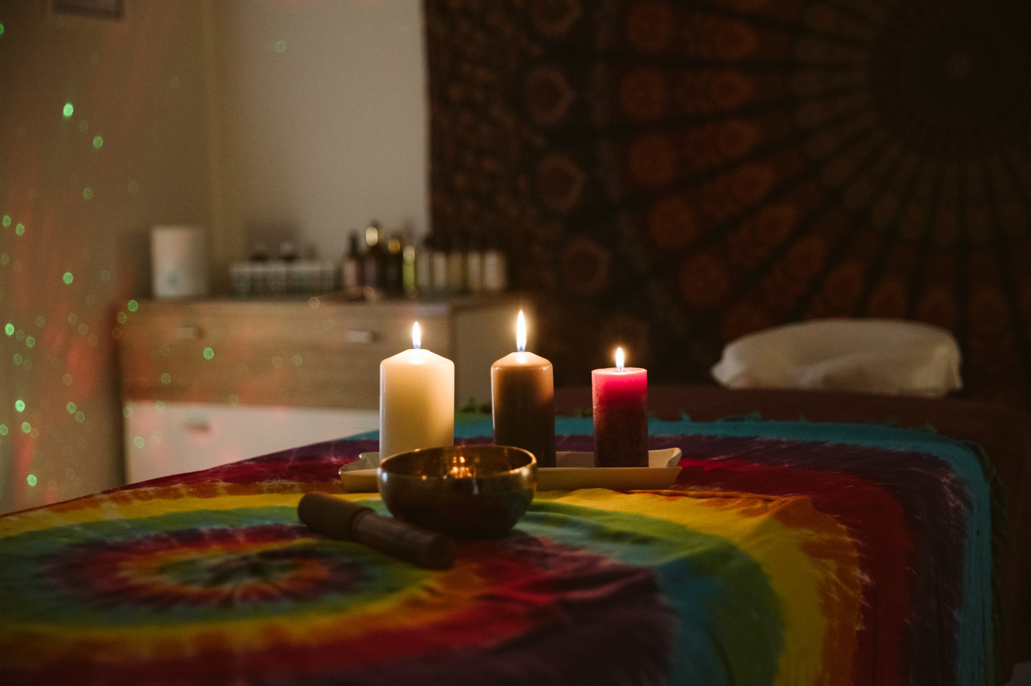 Przytulne wnętrze gabinetu z łóżkiem do zabiegów, na którym leży kolorowy pled w stylu tie-dye, a na nim ustawione trzy zapalone świece o różnych kolorach i miska tybetańska z pałeczką; w tle...