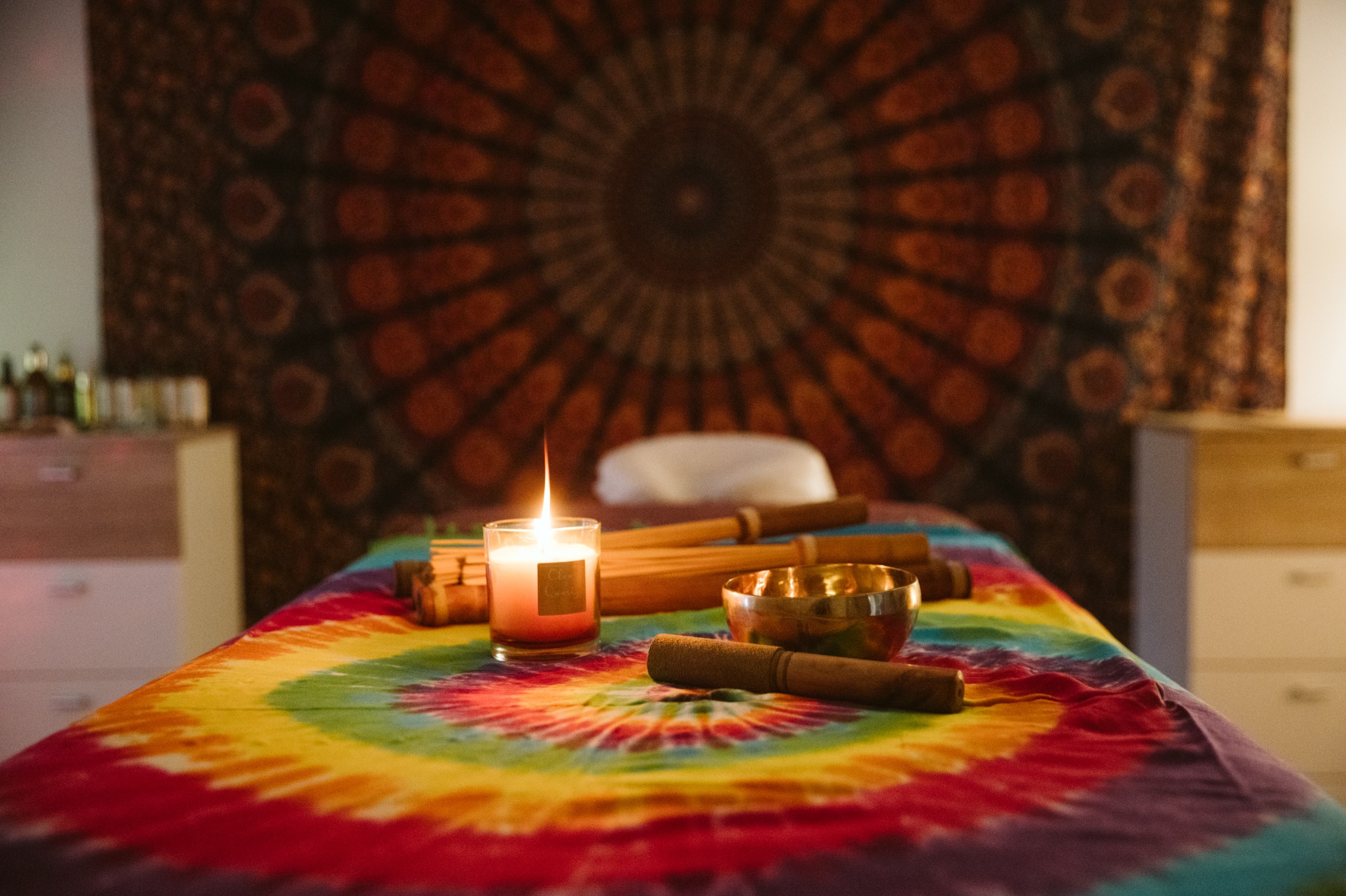 Stół do masażu przykryty kolorową tkaniną w stylu tie-dye, na nim świeca, miseczka dźwiękowa i bambusowe pałeczki, w tle dekoracyjna mandala na ścianie.