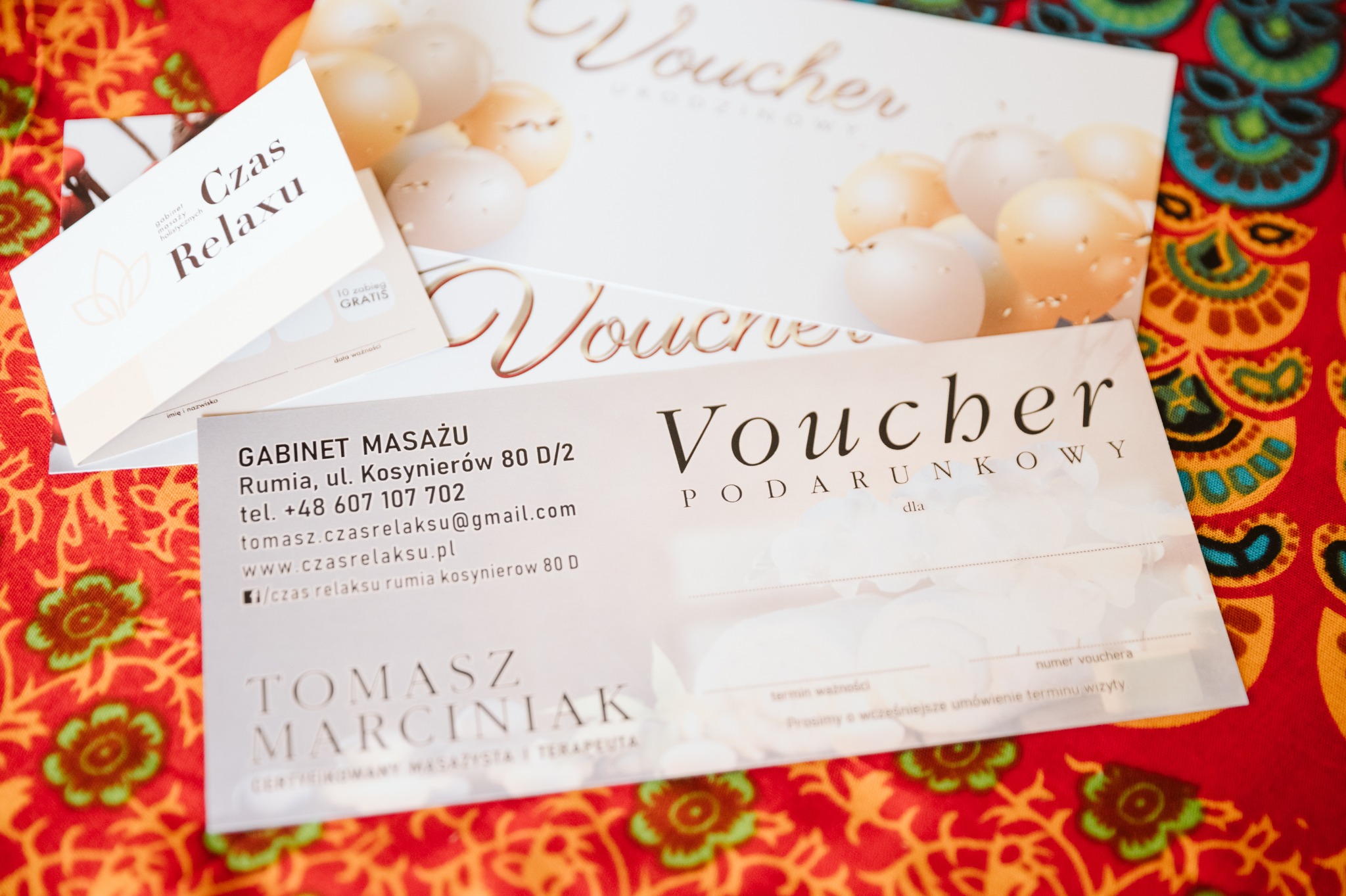 Vouchery podarunkowe i wizytówka gabinetu masażu 'Czas Relaxu' na wzorzystym, czerwonym tle.