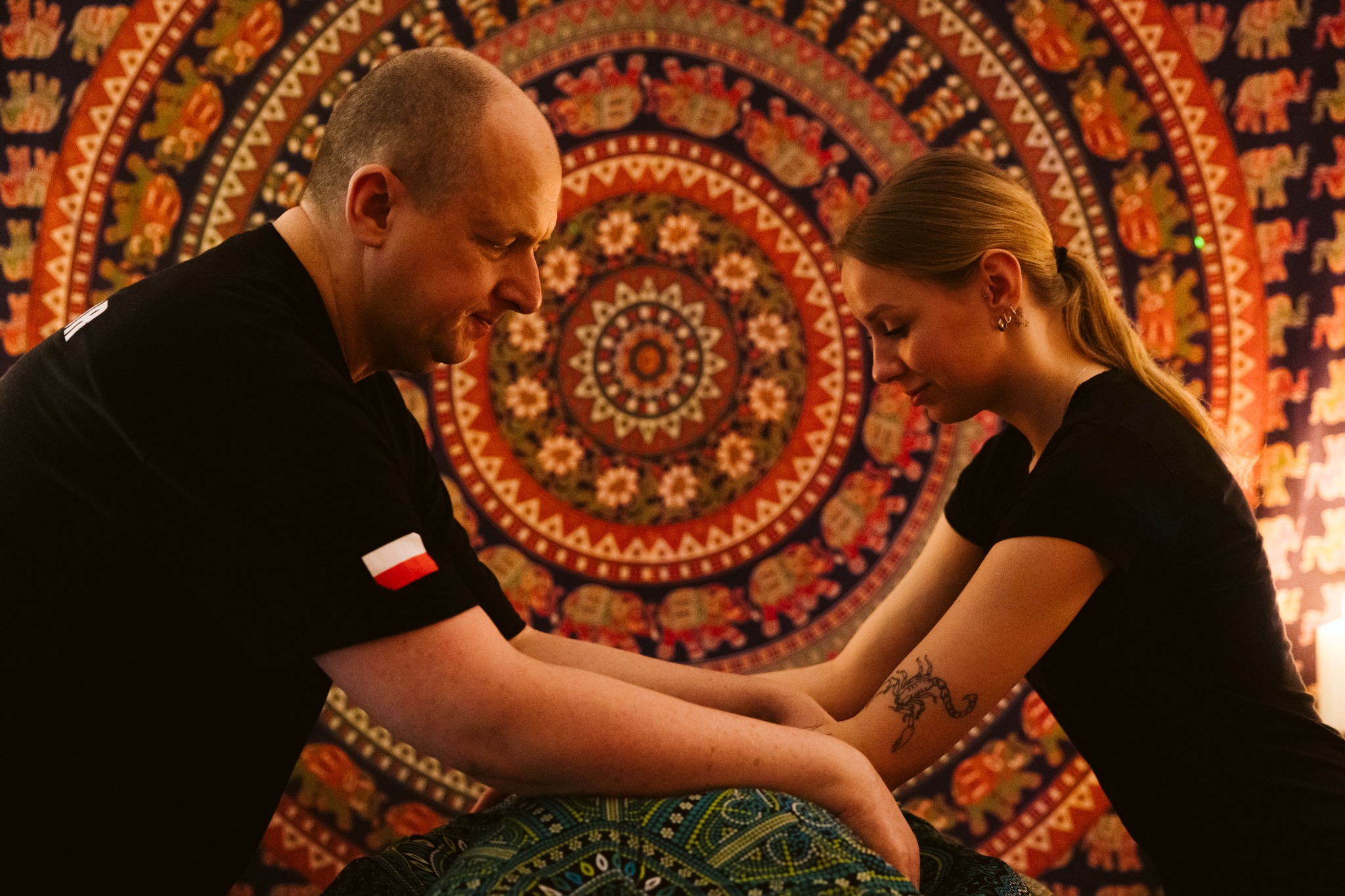 Osoba w czarnej koszulce z flagą Polski wykonuje zabieg na kobiecie z tatuażem skorpiona na ramieniu, na tle wzorzystej mandali.