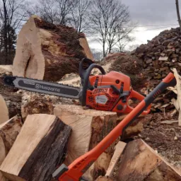 Pomarańczowa pilarka spalinowa Husqvarna 592 XP i rozłupywarka do drewna na stosie świeżo pociętych klocków drewna, w tle stos drewna opałowego i nagie drzewa pod pochmurnym niebem.