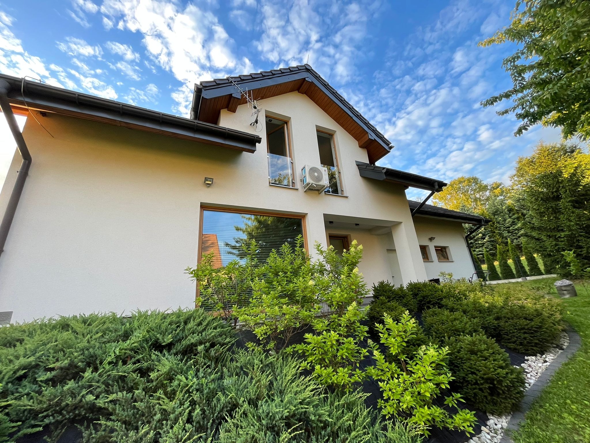 Biały dom jednorodzinny z zamontowaną jednostką klimatyzacji na elewacji, widok z dołu na tle błękitnego nieba z chmurami.