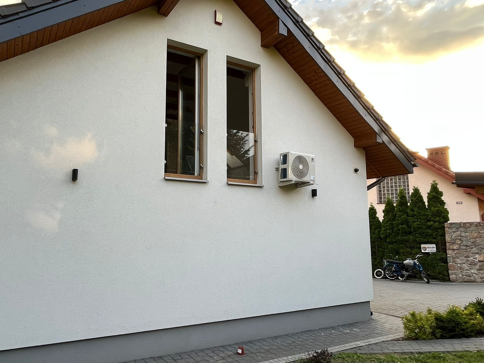 Biała elewacja domu z zamontowaną jednostką zewnętrzną klimatyzacji, widoczne dwa okna z drewnianą ramą, na podjeździe stoi motorower.