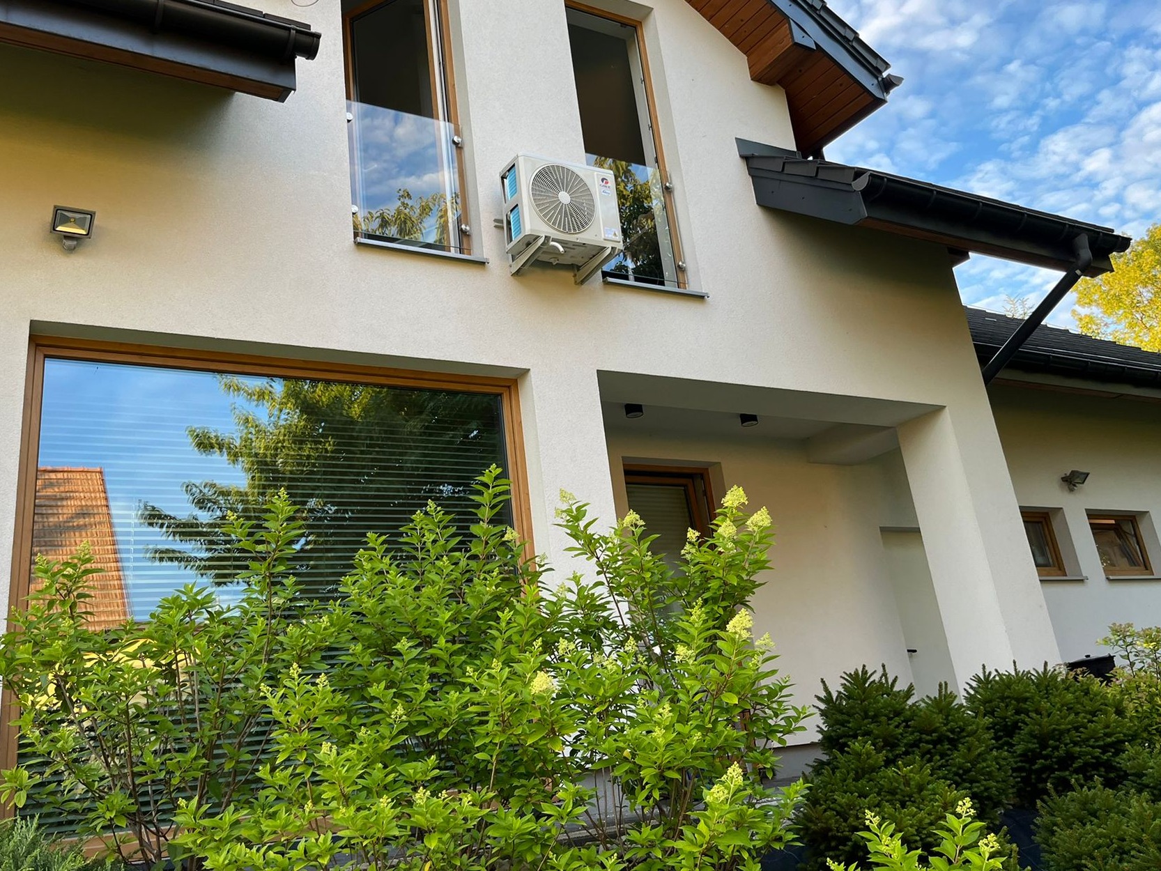 Biała jednostka zewnętrzna klimatyzacji zamontowana na ścianie nowoczesnego domu z dużymi oknami i szklanymi balustradami.