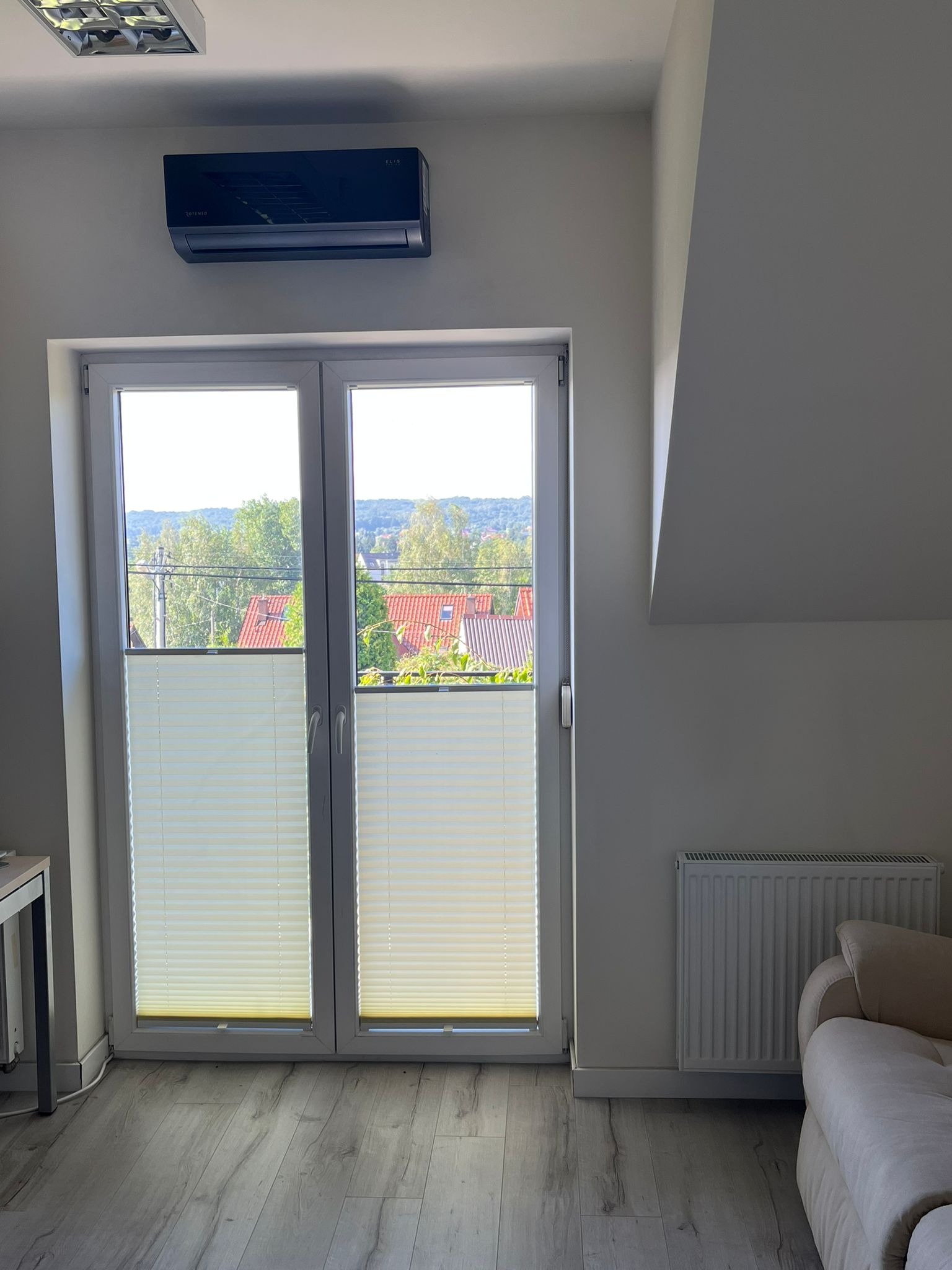 Czarny klimatyzator na ścianie nad białymi drzwiami balkonowymi z roletami plisowanymi, widok na zielone wzgórza za oknem, jasne wnętrze z podłogą imitującą drewno.