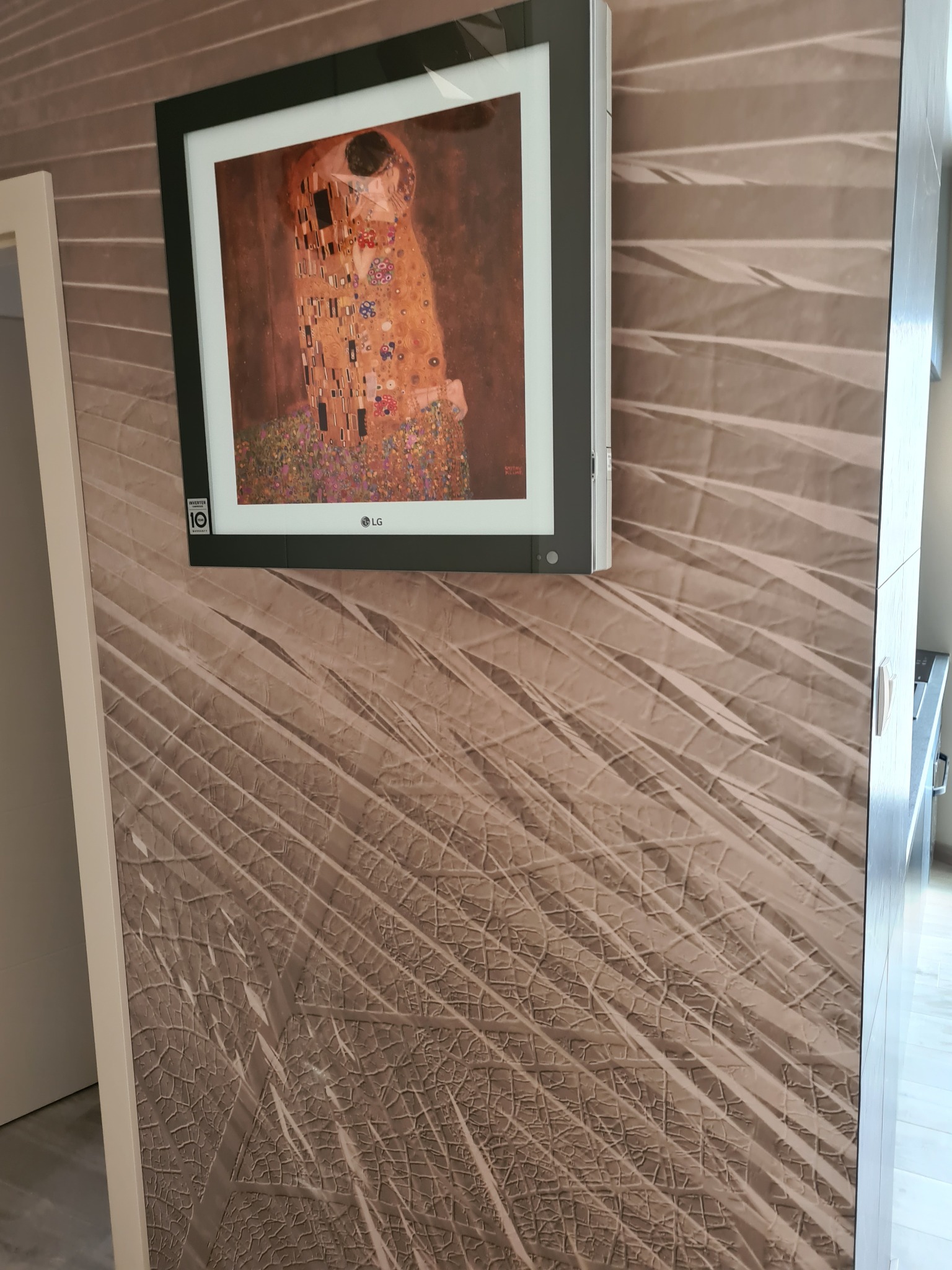 Klimatyzator LG z obrazem Gustava Klimta 'Pocałunek' na ścianie z wzorem liści palmowych.