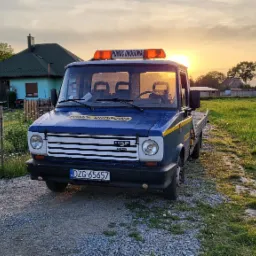 Niebieska laweta DAF z napisem 'Pomoc Drogowa' na dachu, zaparkowana na żwirowej drodze, na tle zielonego pola i zachodzącego słońca.