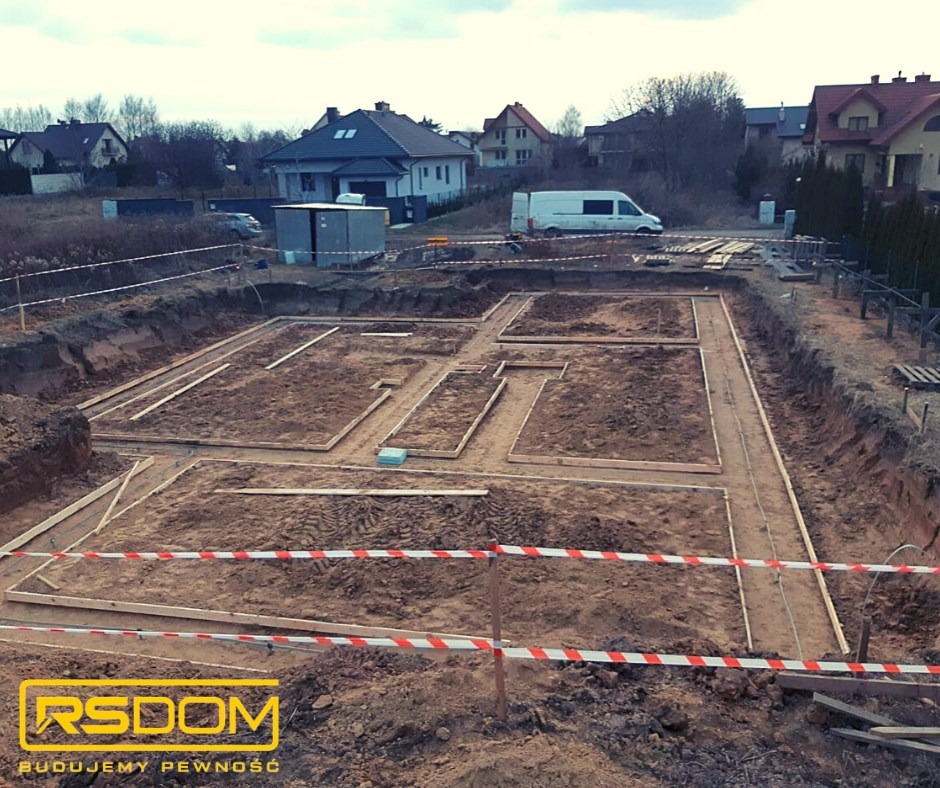 Wykop pod fundamenty domu jednorodzinnego, widoczne szalunki i taśmy ostrzegawcze, w tle zabudowa mieszkalna i bus.