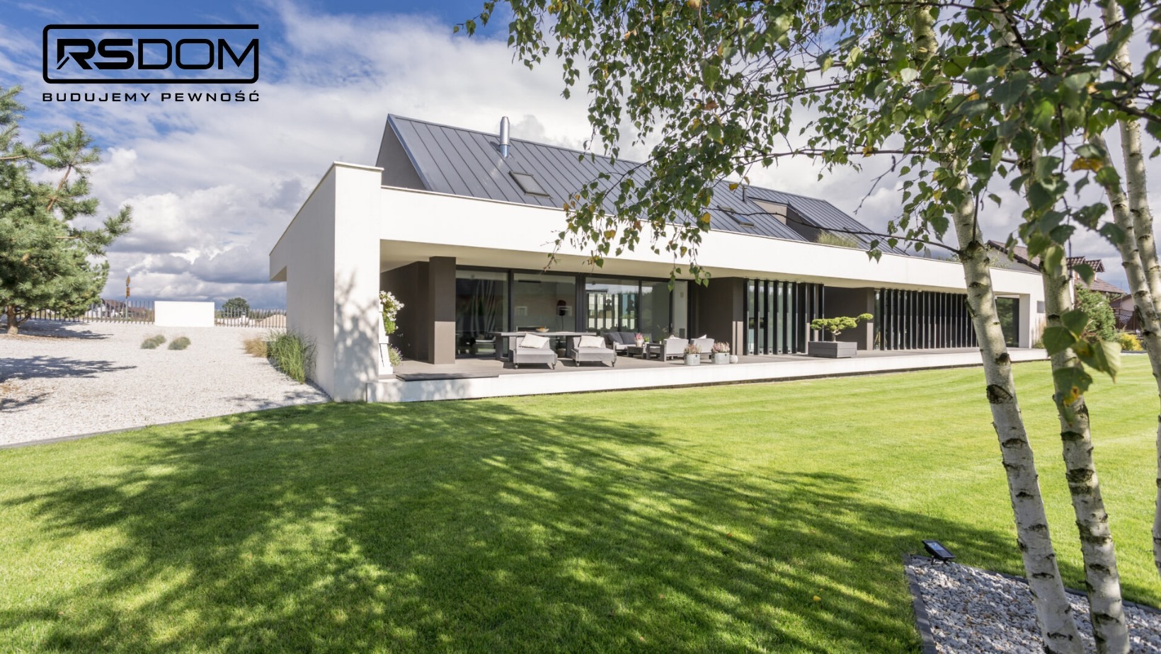 Nowoczesny dom jednorodzinny z płaskim dachem i dużym tarasem, widok z ogrodu z drzewami brzozy na pierwszym planie i logo firmy budowlanej w lewym górnym rogu.