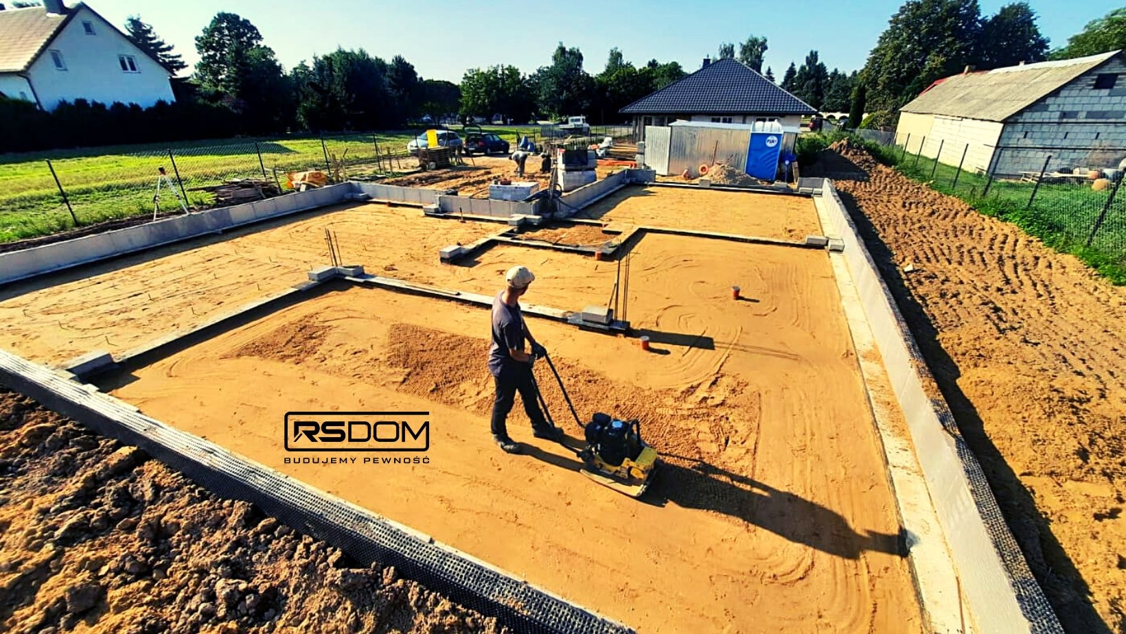 Prace ziemne przy budowie fundamentów domu jednorodzinnego: ubijanie piasku zagęszczarką wewnątrz betonowych szalunków, widoczne zbrojenia i przygotowany teren pod wylewkę.