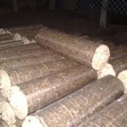 Stos równo ułożonych, cylindrycznych brykietów opałowych w magazynie, widoczne charakterystyczne włókna drewna.