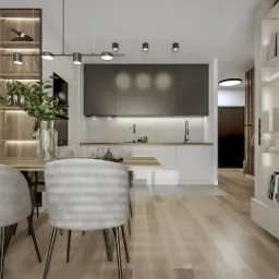 Jasne wnętrze mieszkania z drewnianym stołem i krzesłami na pierwszym planie, w tle minimalistyczna kuchnia z szarymi szafkami i białą wyspą, po prawej stronie regał z podświetlanymi półkami...