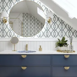 Nowoczesna łazienka na poddaszu z niebieską szafką pod umywalką, okrągłym lustrem w złotej ramie, wzorzystą tapetą i oknem dachowym.