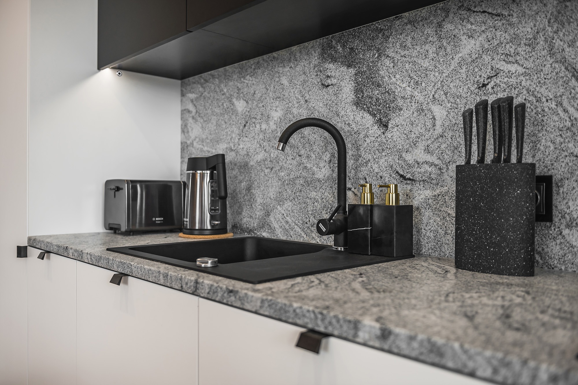 Nowoczesna kuchnia z czarnym zlewem, baterią i akcesoriami na blacie z szarego kamienia, uzupełniona o czajnik, toster i blok z nożami, podświetlona punktowym światłem.