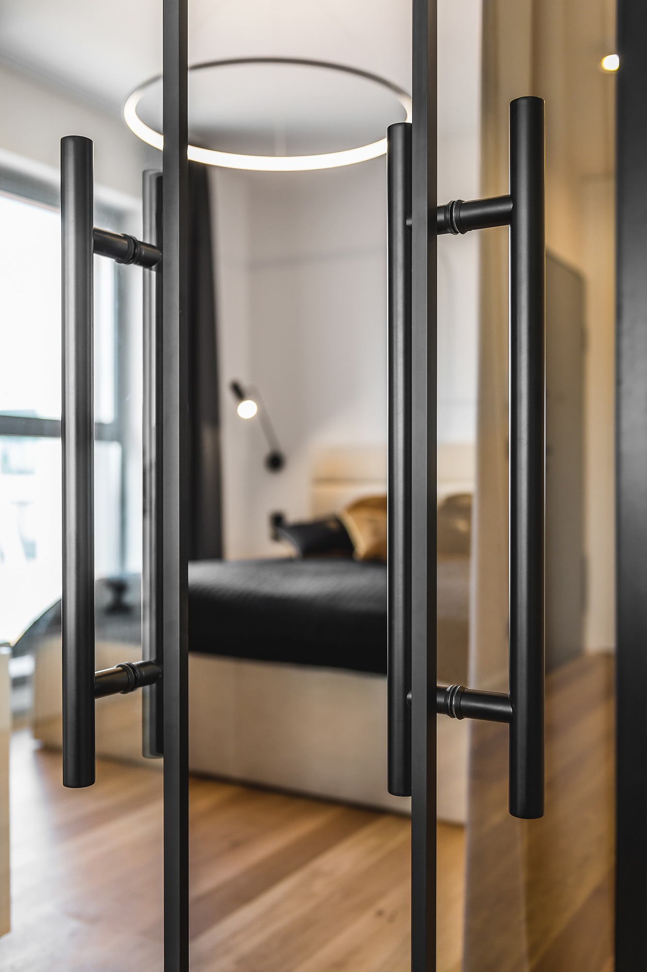 Szklane drzwi z czarnymi, metalowymi uchwytami na tle sypialni z drewnianą podłogą, widoczna okrągła lampa sufitowa i łóżko z czarną pościelą.