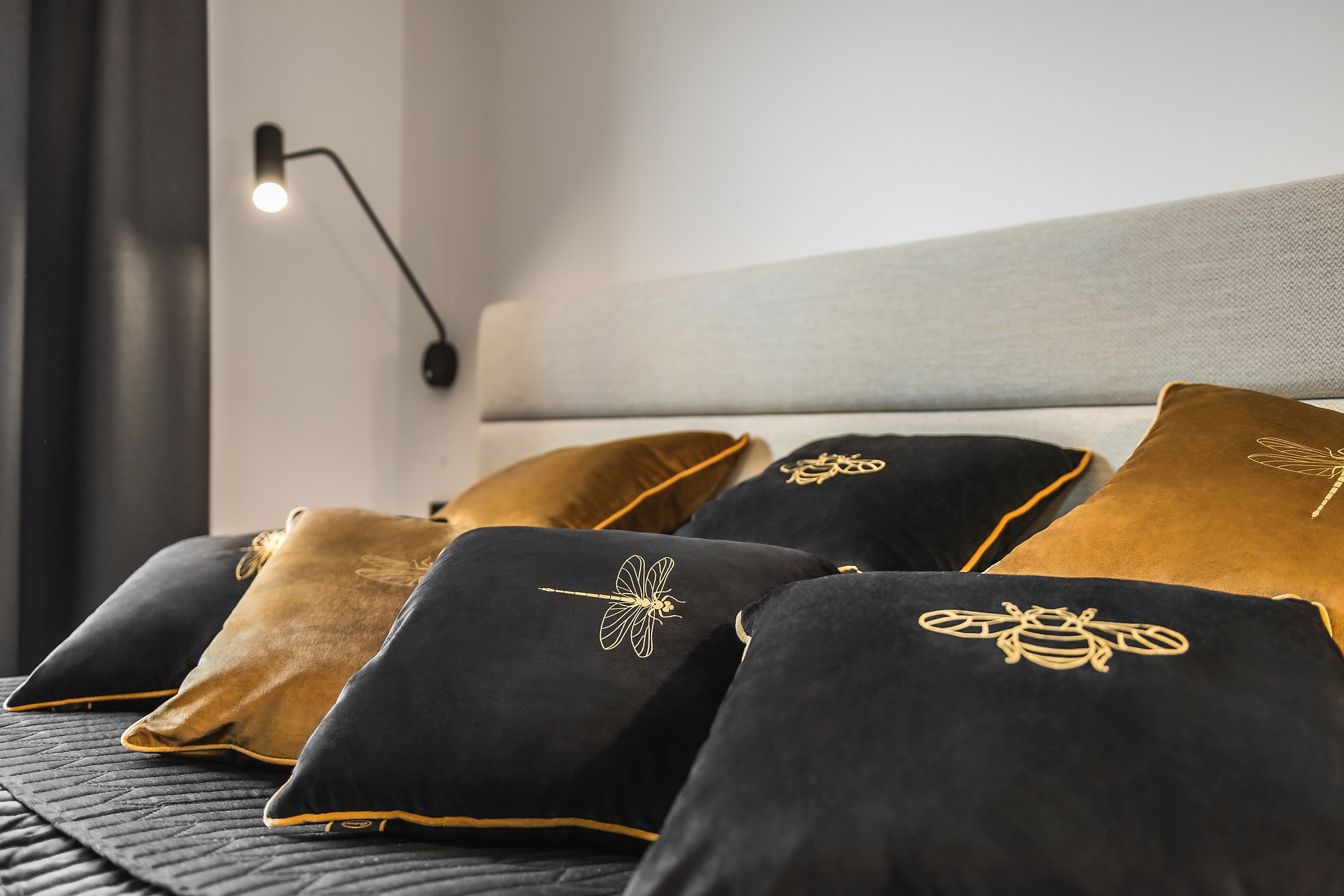 Dekoracyjne poduszki w odcieniach złota i czerni z haftowanymi motywami owadów, ułożone na łóżku z szarą tapicerką i czarną narzutą, oświetlone przez nowoczesną lampę ścienną.