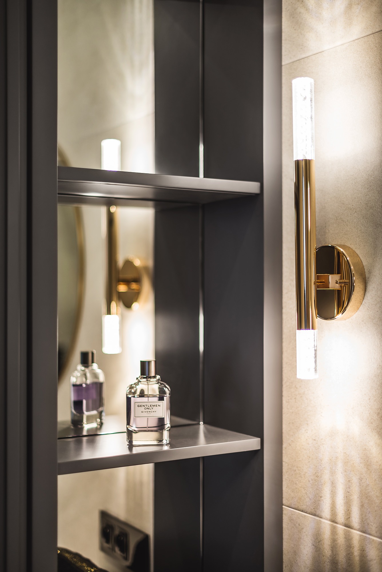 Szara szafka łazienkowa z lustrzanymi plecami, oświetlona złotą lampą ścienną, na półkach flakony perfum.