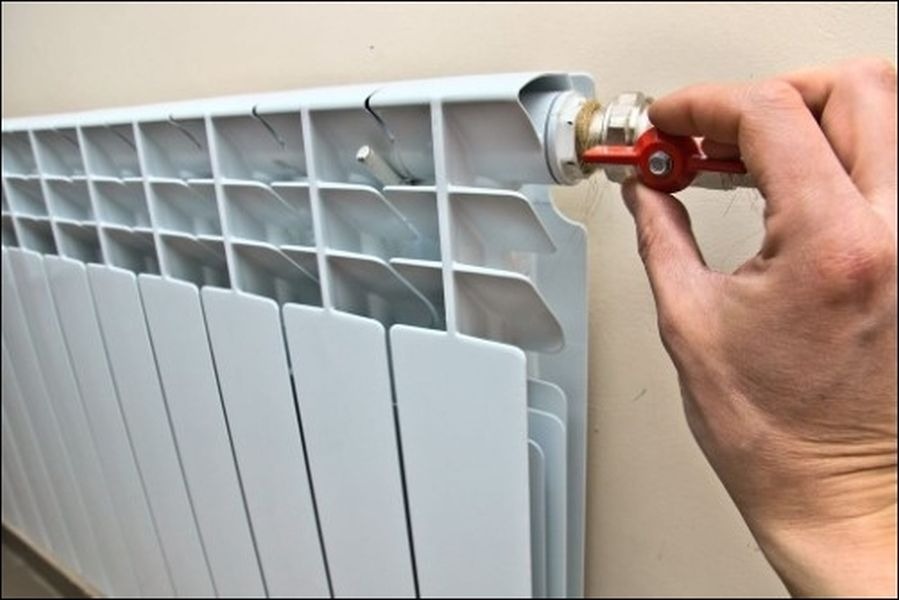 Biały grzejnik aluminiowy, regulowany zaworem z czerwoną rączką przez dłoń, widok z bliska.