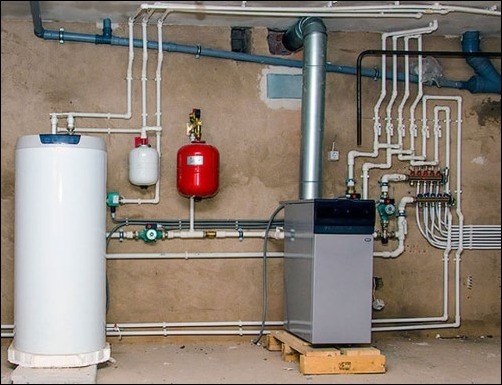 Zestaw instalacji centralnego ogrzewania w piwnicy: biały zasobnik ciepłej wody, szary piec na drewnianej palecie, czerwony zbiornik wyrównawczy, liczne rury z tworzywa sztucznego i metalu.