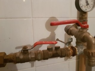 Stare, skorodowane zawory z czerwonymi rączkami i manometrem na tle białych, kwadratowych płytek. Widoczne ślady rdzy i osadu na metalowych elementach instalacji.