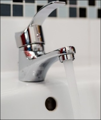 Błyszcząca chromowana bateria umywalkowa z wypływającą wodą na tle białej umywalki i mozaiki ściennej.