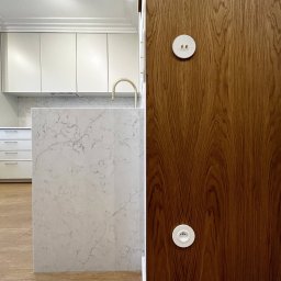 Coren Studio Design - Kuchenna wyspa z marmurowym blatem i drewniana ściana z włącznikami w stylu retro, w jasnym wnętrzu. Widok na zabudowę kuchenną w tle.