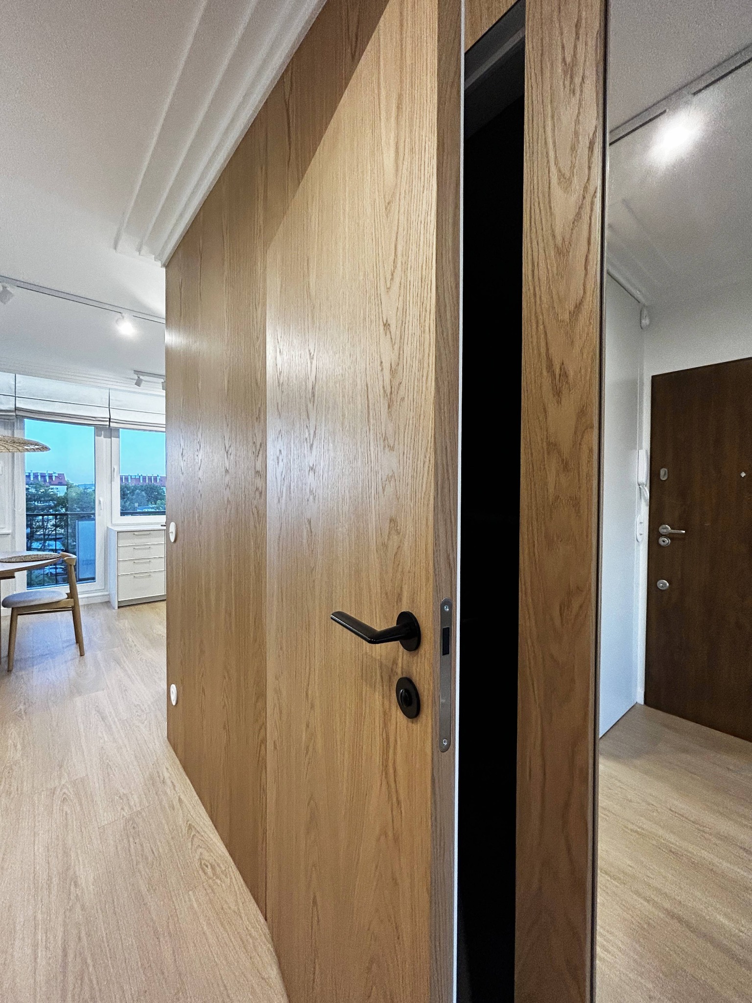 Nowoczesne drzwi wewnętrzne z drewnianą okleiną, czarny uchwyt, widok na odnowione mieszkanie z balkonem w tle. Gdańsk Brzeźno, rynek wtórny.