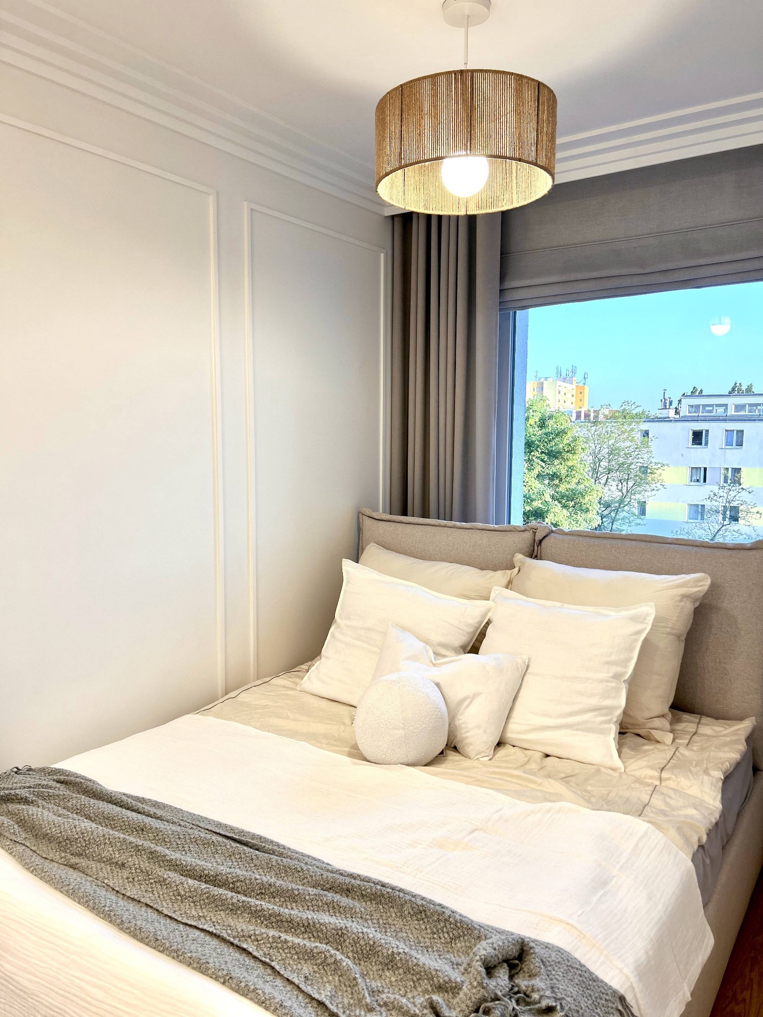 Jasna sypialnia z łóżkiem, beżową narzutą i dekoracyjnymi poduszkami. Okno z widokiem na miasto. Lampa z naturalnego materiału. Ściana z dekoracyjnymi listwami.