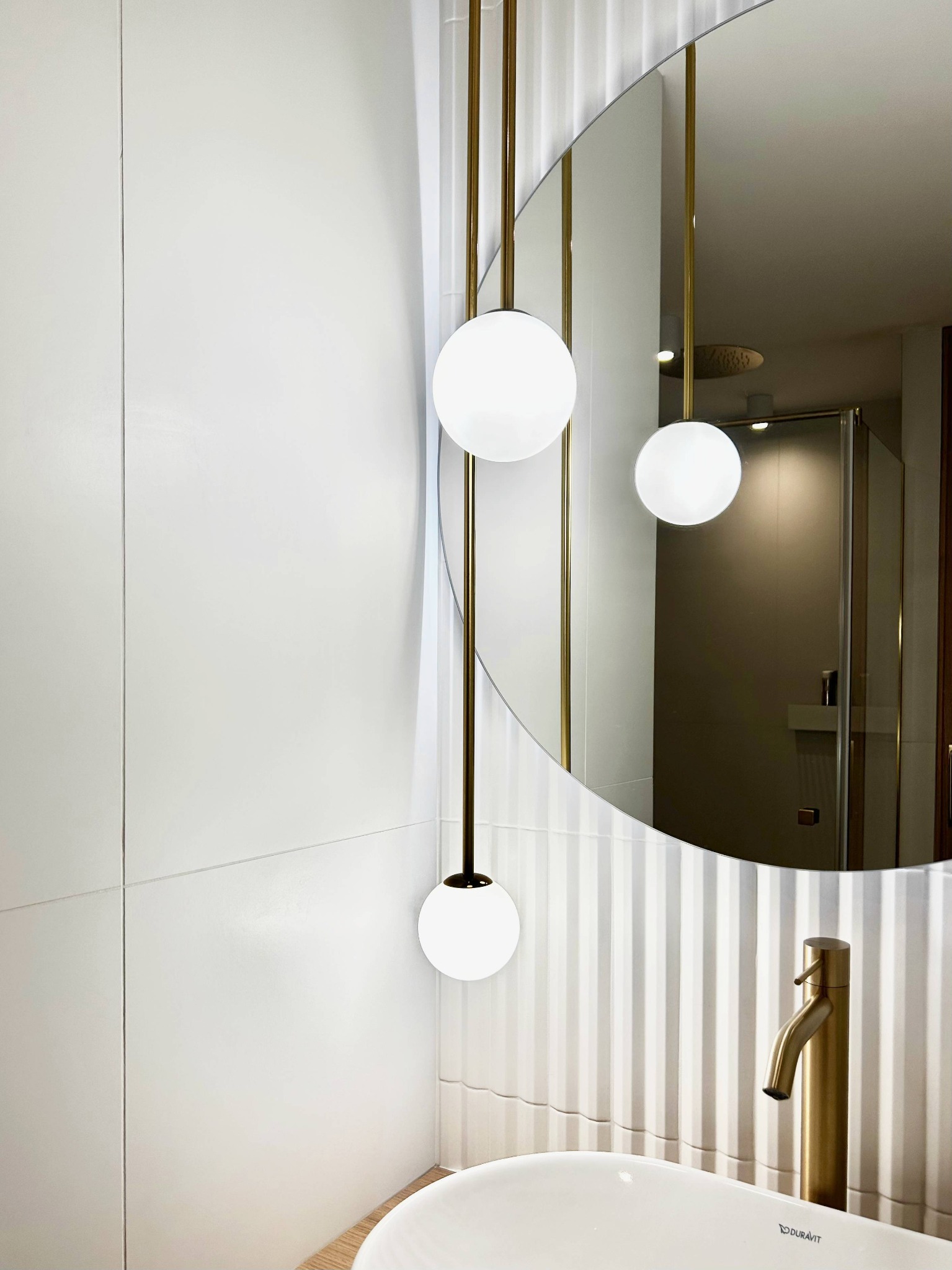Nowoczesna łazienka z okrągłym lustrem, złotą armaturą i lampami kulami. Białe płytki ścienne z subtelnym wzorem. Widoczna kabina prysznicowa w odbiciu lustra.