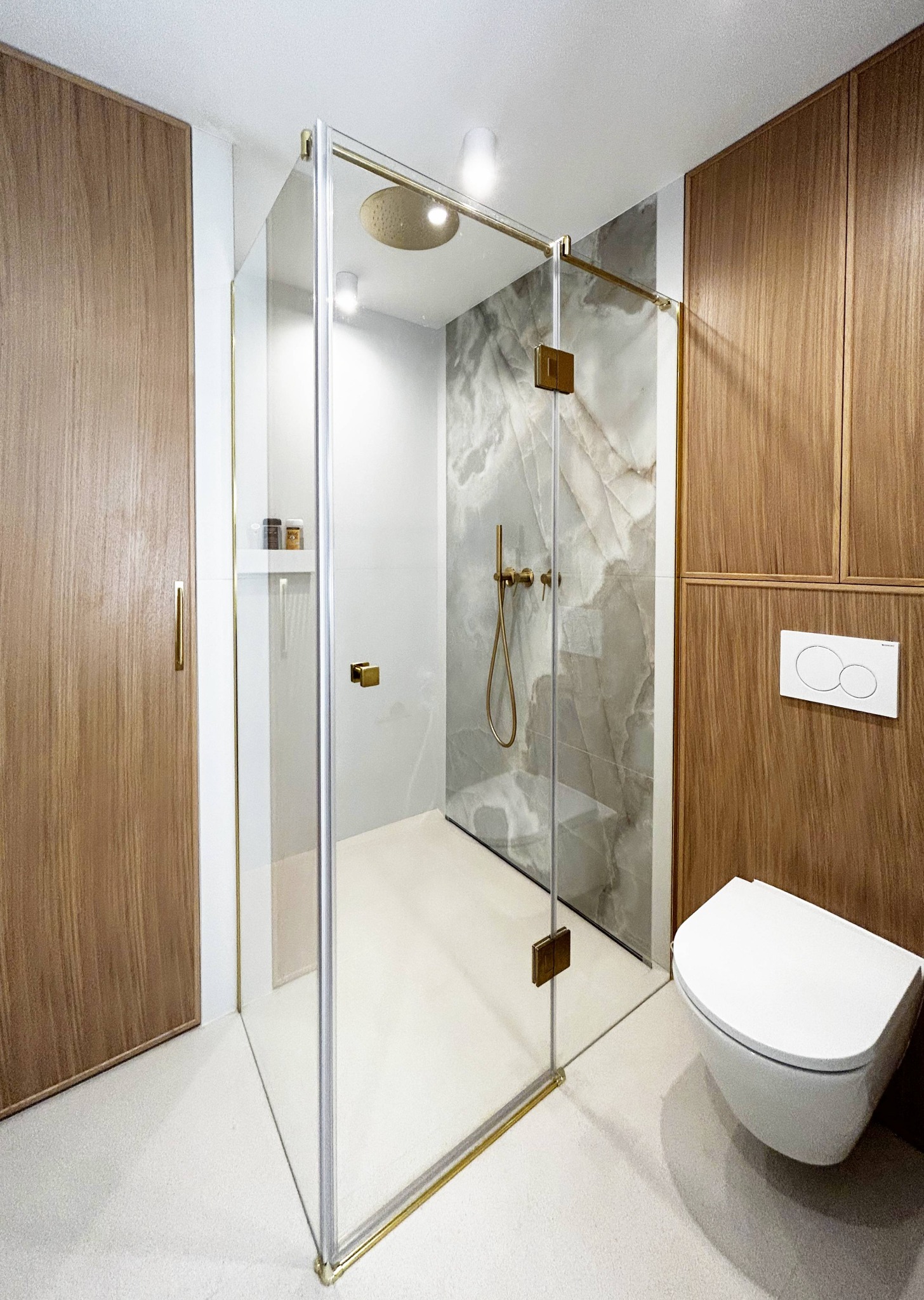 Nowoczesna łazienka z kabiną prysznicową typu walk-in, wykończoną złotymi detalami. Na ścianie marmurkowy wzór. Obok podwieszana toaleta i drewniane szafki.