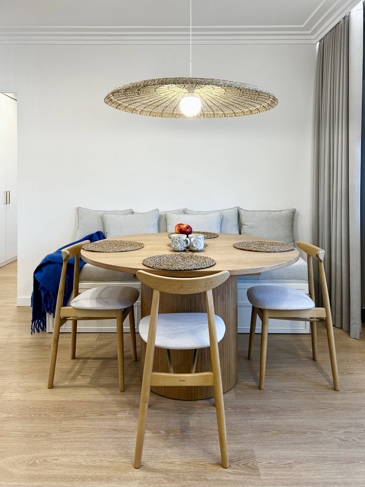 Jasna jadalnia z okrągłym stołem, drewnianymi krzesłami i lampą z abażurem z naturalnego materiału. W tle wnęka z siedziskiem i poduszkami oraz zasłony.