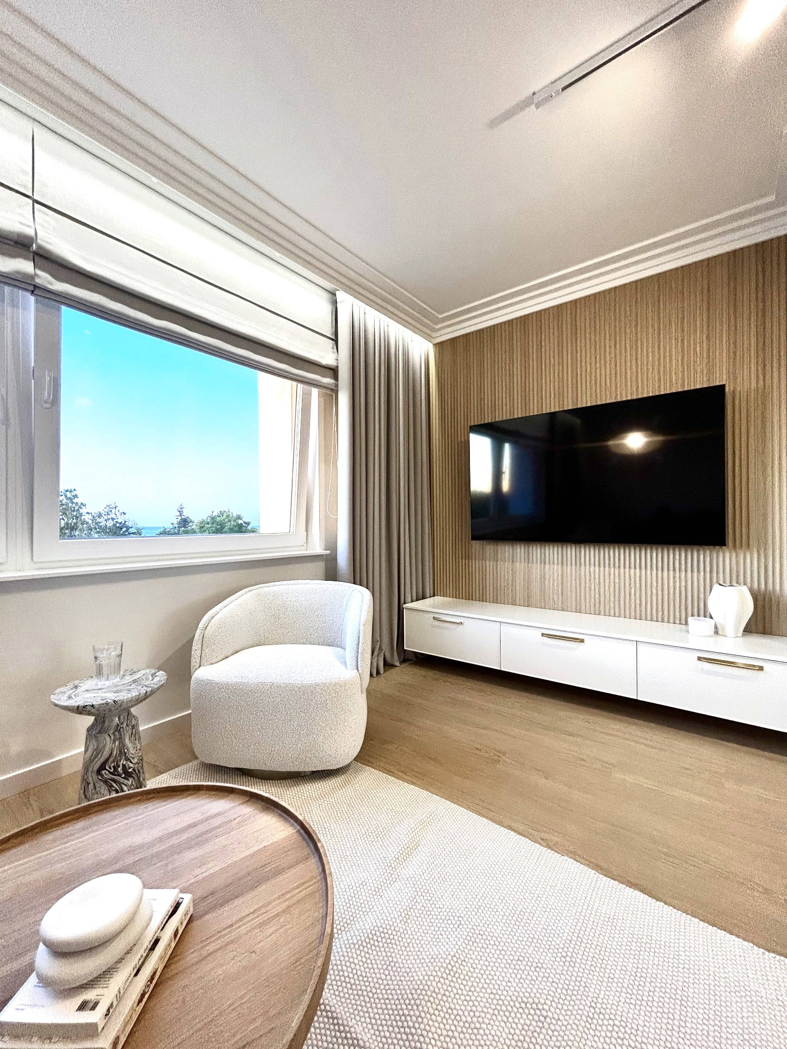 Jasne wnętrze salonu z widokiem na morze: kremowy fotel, stolik marmurowy, drewniany stolik kawowy i ściana z telewizorem. Minimalistyczny design z akcentami natury.