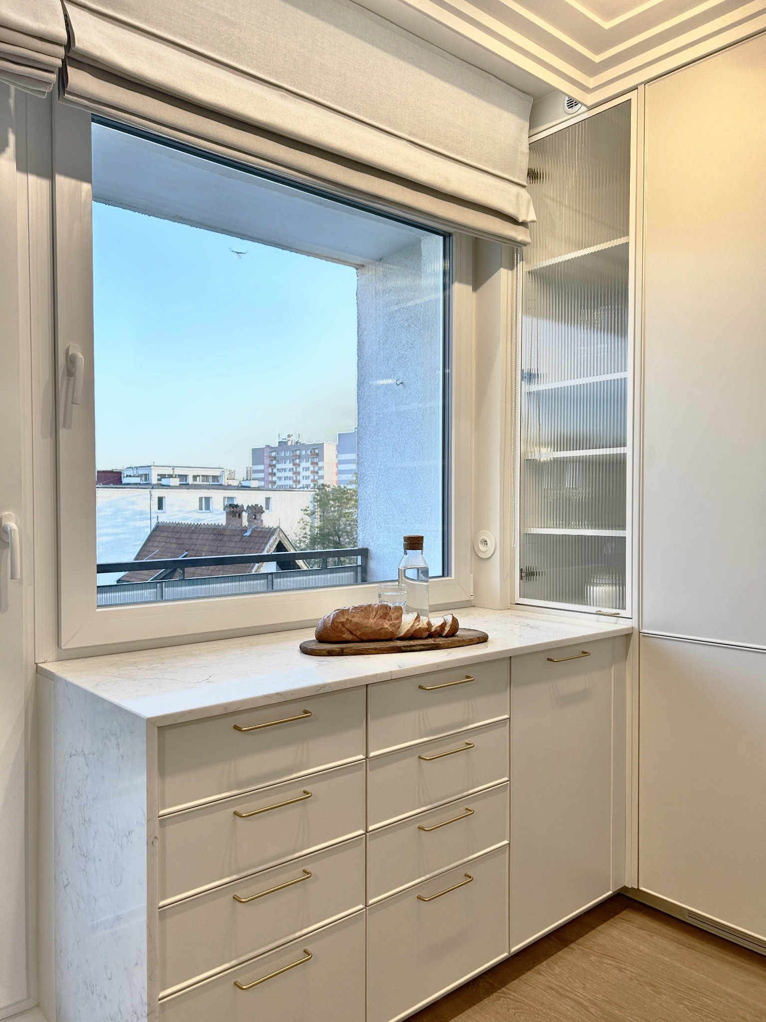 Jasna kuchnia z marmurowym blatem, na którym leży chleb i butelka wody. Okno z roletą rzymską, widok na miasto. Ryflowane szklane drzwi szafki.