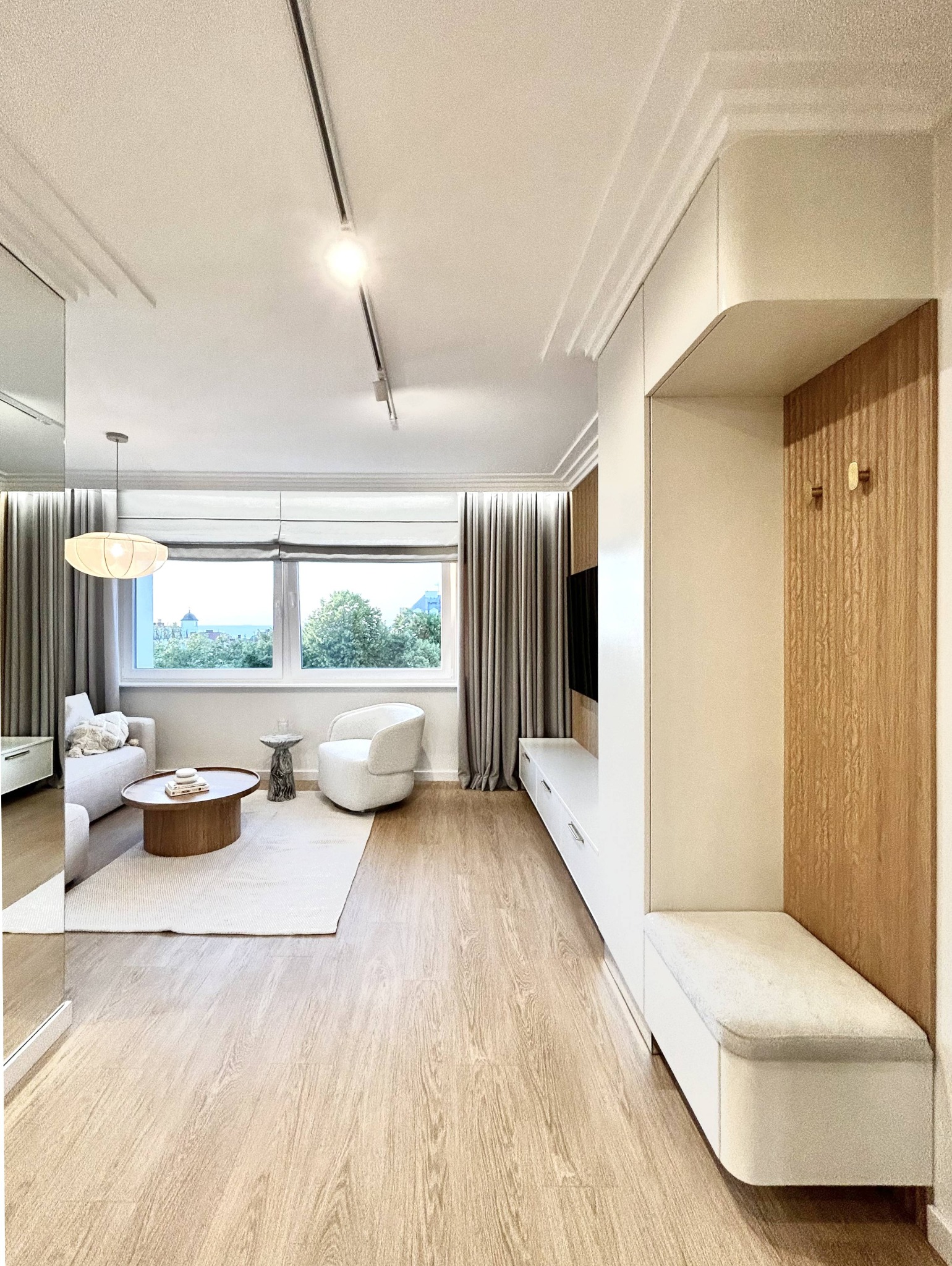 Jasne wnętrze pokoju dziennego z drewnianą podłogą i elementami w stylu skandynawskim. Widok na okno z zielenią i fragment zabudowy z siedziskiem.