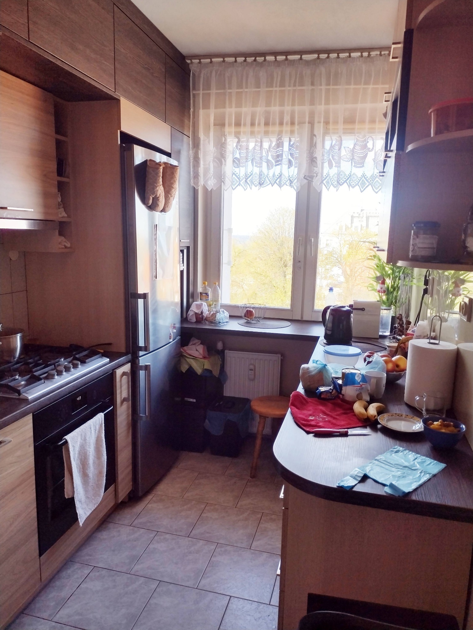 Kuchnia w bloku przed aranżacją: widok na okno z firanką, zabudowa kuchenna z brązowymi frontami, na blacie kuchennym naczynia i produkty spożywcze.