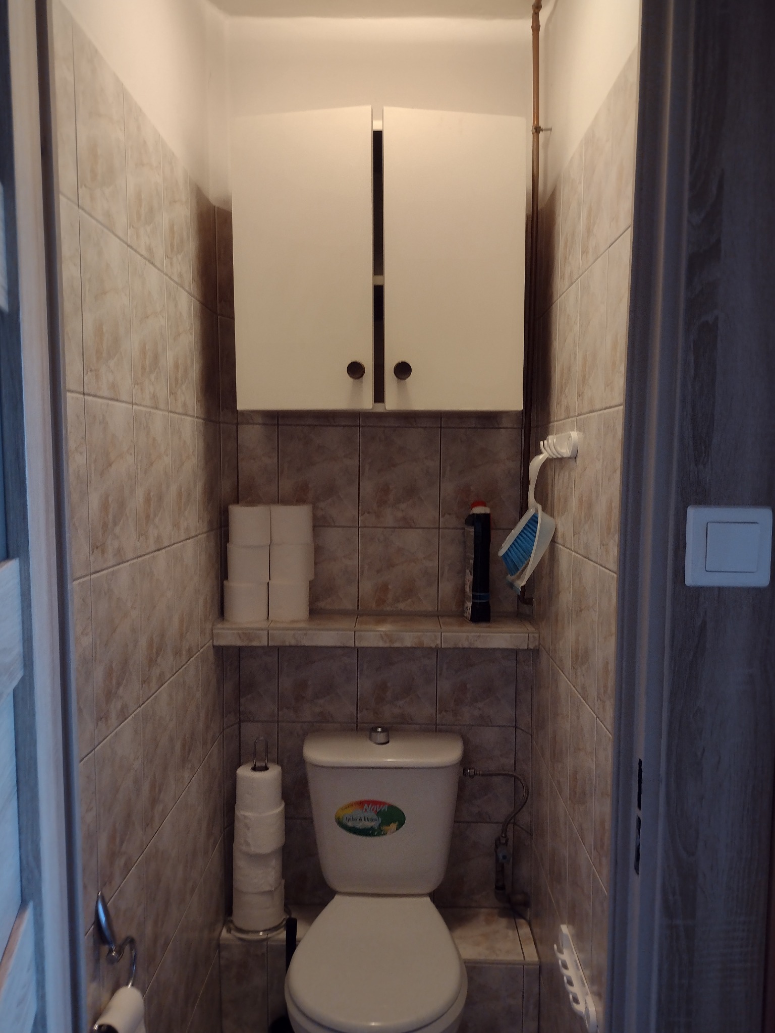 Mała łazienka w beżowych płytkach z toaletą i szafką wiszącą. Widoczne pionowe rury miedziane. Zdjęcie z projektu w Gdańsku Brzeźnie, rynek wtórny, stan PRZED.