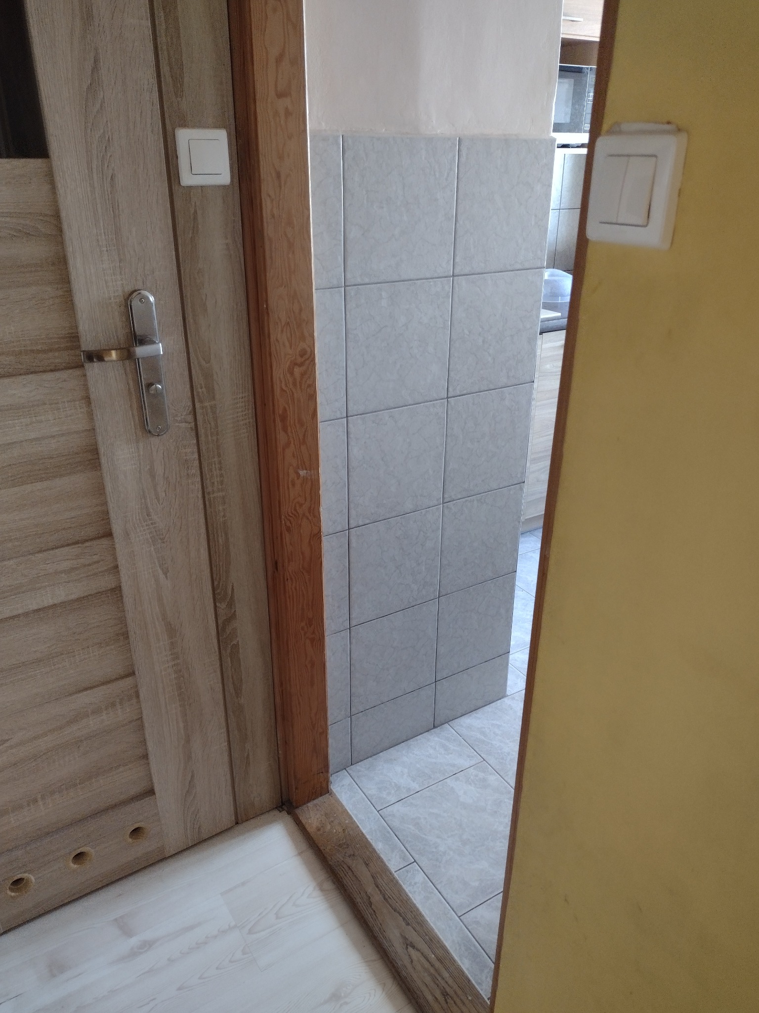 Fragment łazienki z widocznymi płytkami na ścianie i podłodze, drzwi z klamką oraz włącznik światła. Zdjęcie 'przed' remontem, z perspektywy wejścia do pomieszczenia.