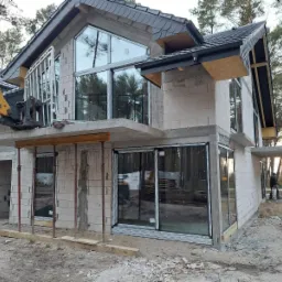 Montaż okien na budowie domu jednorodzinnego; widoczny podnośnik do szyb i surowy stan elewacji z betonu komórkowego.