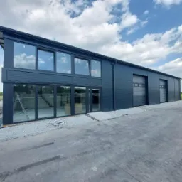 Nowoczesny budynek magazynowy z antracytową elewacją, dużymi oknami i bramami garażowymi segmentowymi, częściowo wykończony teren wokół budynku.