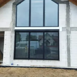 Nowoczesny dom w trakcie budowy z zamontowanymi oknami z czarnymi ramami i opuszczonymi czarnymi roletami zewnętrznymi, widoczne surowe ściany z betonu komórkowego.