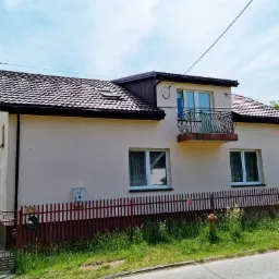 Dom jednorodzinny z brązowym dachem z dachówki ceramicznej, oknem dachowym i balkonem z kutą balustradą, otoczony czerwonym płotem sztachetowym.