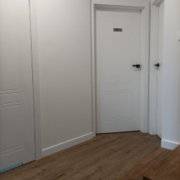 Pośrednictwo Możarowski - Korytarz biurowy z trzema białymi drzwiami, brązową podłogą imitującą drewno i czarnymi klamkami. Na środkowych drzwiach tabliczka z napisem 'DYREKTOR'.