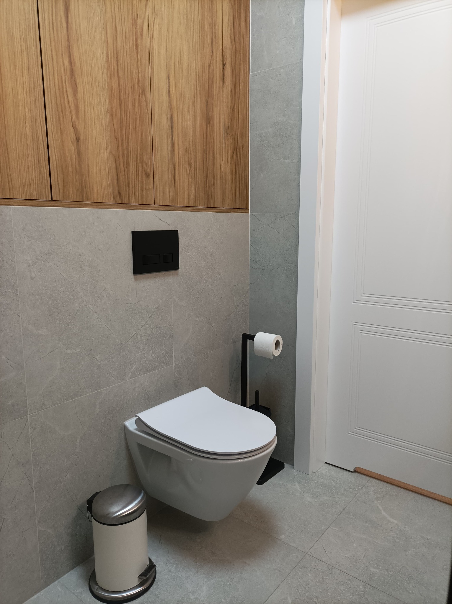 Nowoczesna toaleta w łazience z szarą płytką imitującą marmur, podwieszana miska WC, drewniane szafki wiszące i białe drzwi.