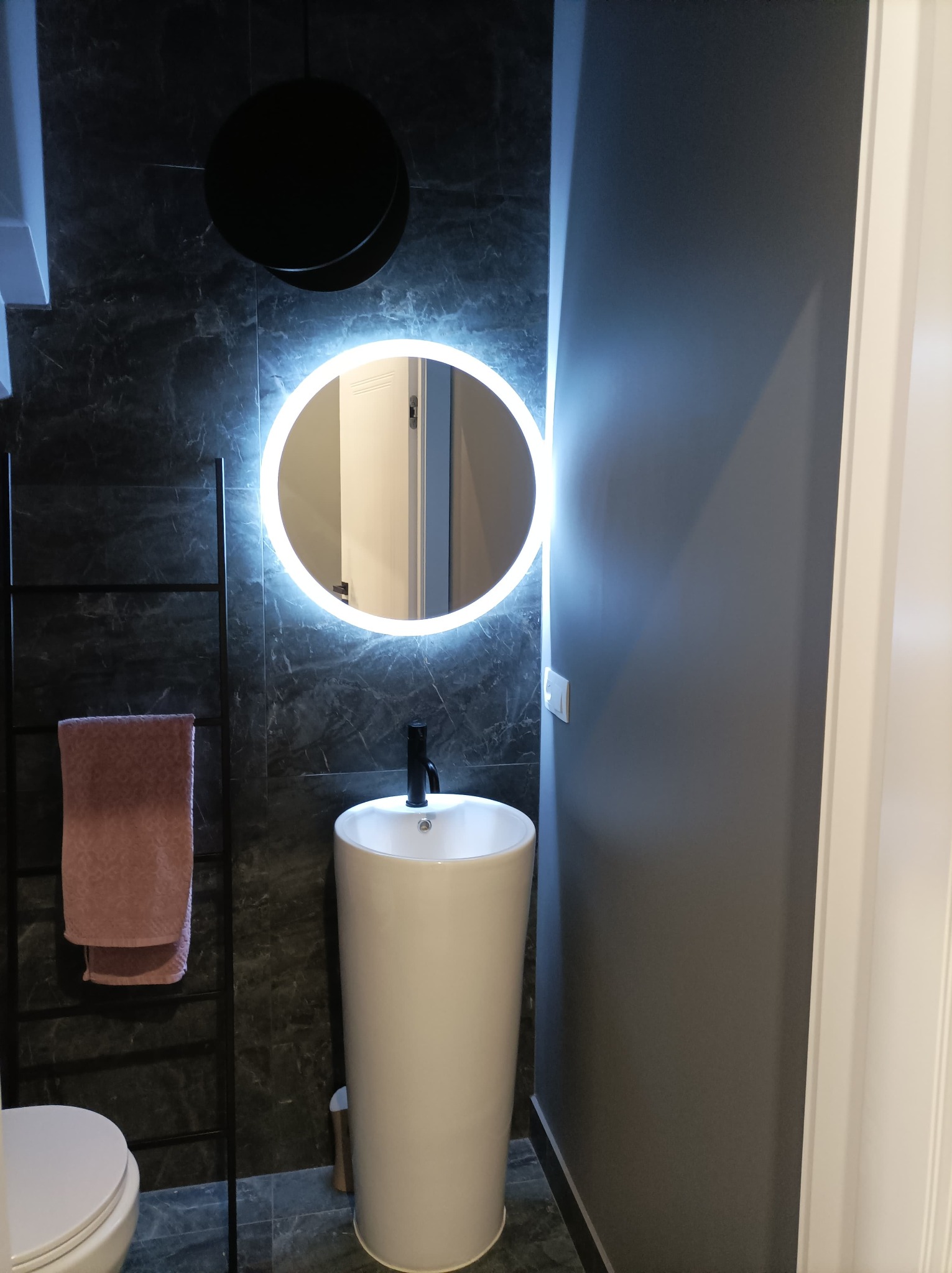 Nowoczesna łazienka z okrągłym lustrem LED, wolnostojącą umywalką i czarną drabinką na ręczniki na tle ciemnej marmurowej ściany.